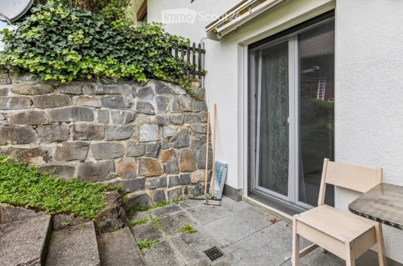 Small outdoor patio area with a stone wall, wooden chair, small table, broom, and a sliding glass door leading to the house.