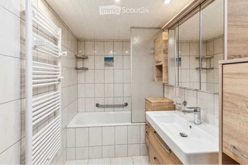 Modern bathroom with a bathtub, sink, mirror, and wooden cabinet. Features tiled walls and a white radiator on the left side.