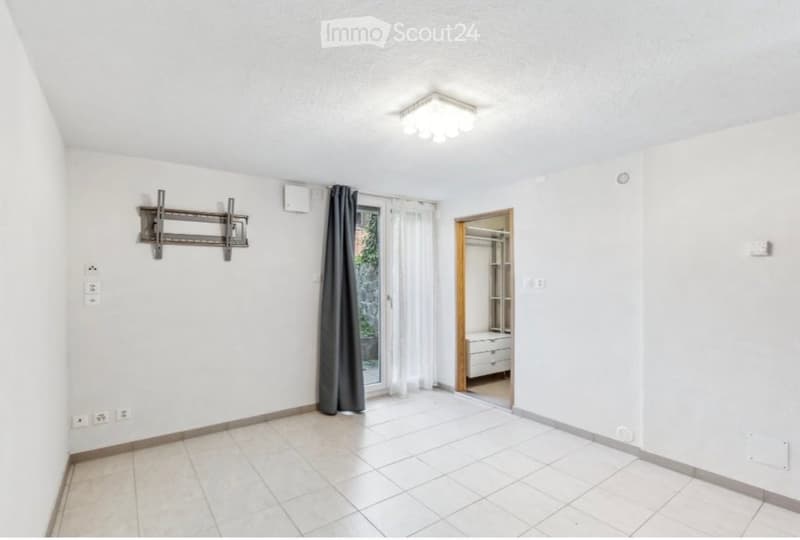 Empty room with white walls, tiled floor, and a sliding glass door leading to a balcony. A TV mount is attached to the wall, and a small closet with drawers is visible in the adjacent space.