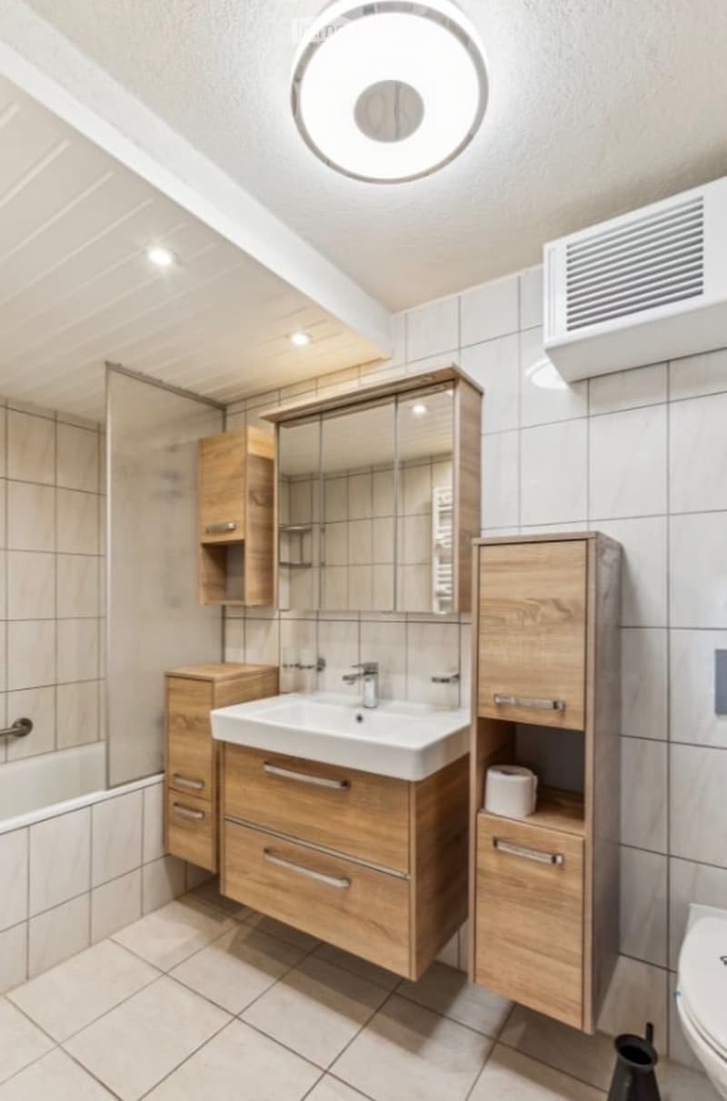 Modern bathroom with wooden cabinets, a white sink, a large mirror, and a tiled shower area.