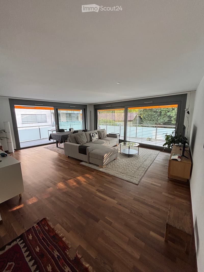 Modern living room with wooden flooring, large windows, grey sectional sofa, coffee table, ottoman, side table, TV, and plants.