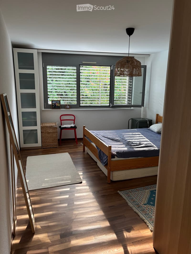 Bright bedroom with wooden flooring, large windows with blinds, a wooden bed with blue bedding, a red chair, a wicker basket, a nightstand with a lamp, and a patterned rug.