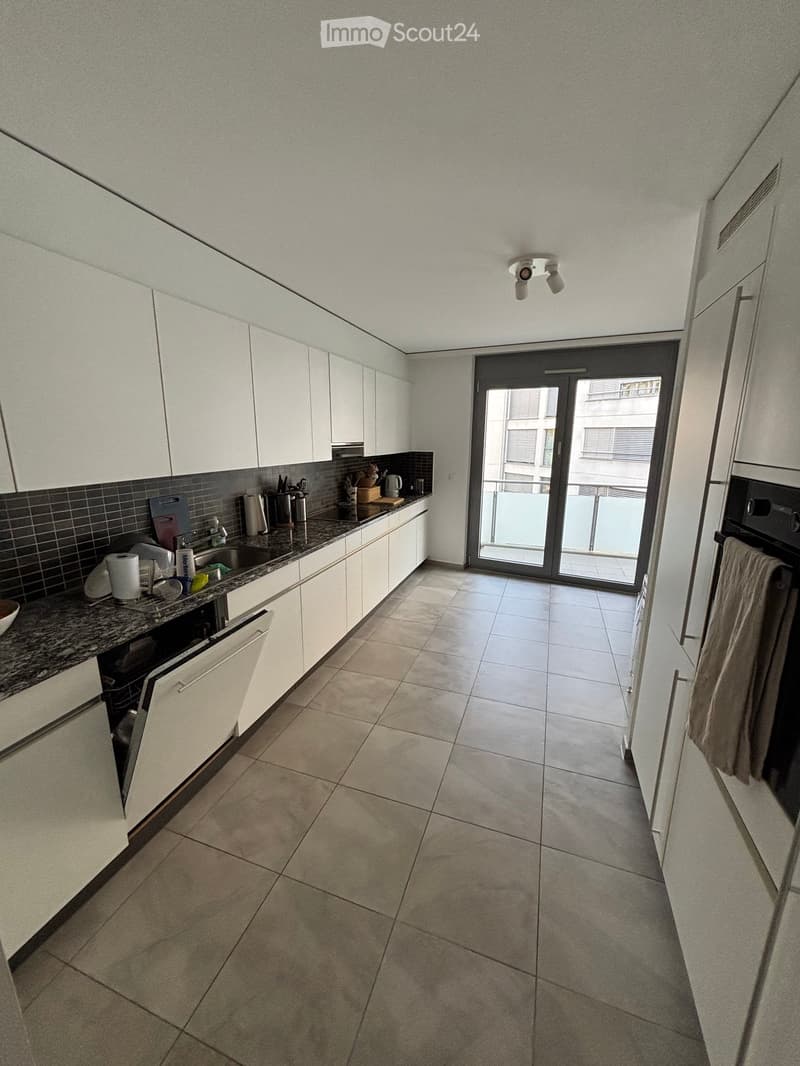 Modern kitchen with white cabinets, granite countertop, and tiled floor. Features a sink, dishwasher, and oven, with a sliding glass door leading to a balcony.