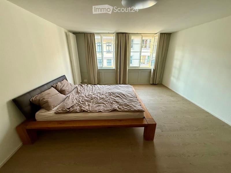 A modern bedroom with a wooden bed, beige bedding, and large windows with curtains. The room features light-colored walls and a wooden floor.