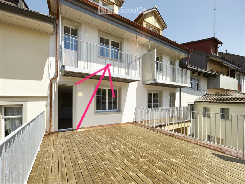 Wooden balcony with metal railings, overlooking a multi-story residential building with multiple balconies and windows.