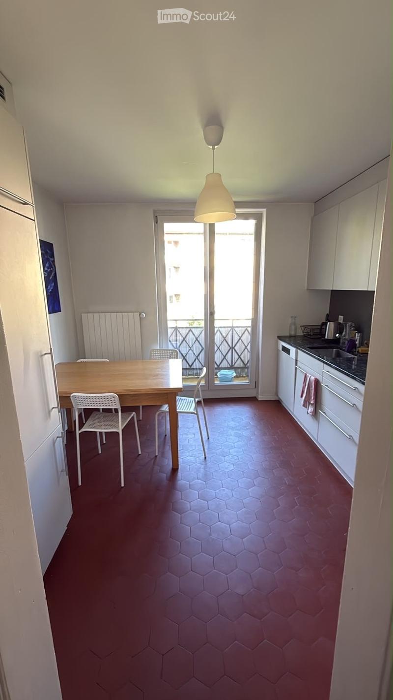A modern, open-plan kitchen and dining area with a hexagonal red tile floor, white cabinets, a wooden dining table with four white chairs, and a hanging pendant light.