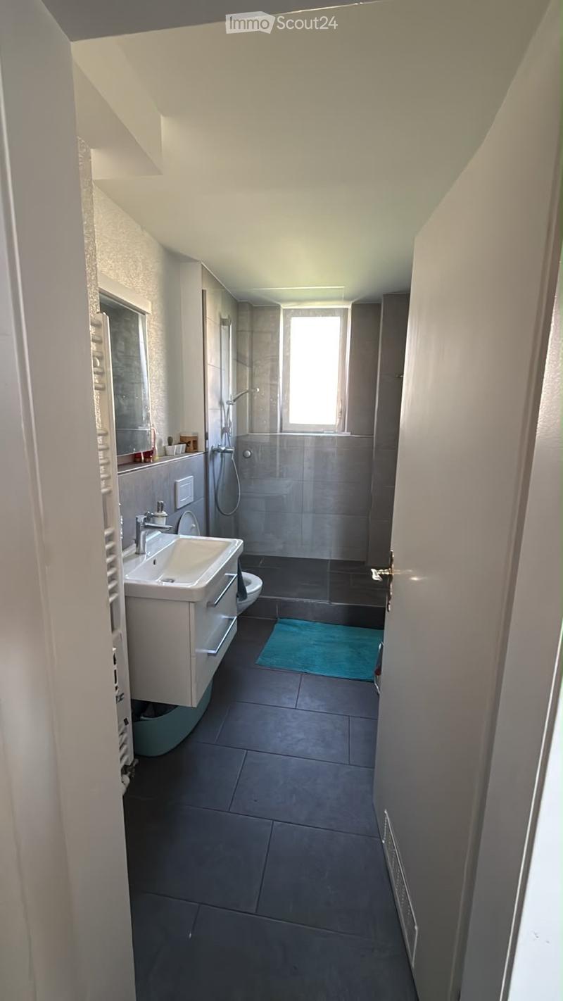 Modern bathroom with a white sink, mirror, toilet, and a shower area with a window. The floor is tiled in dark gray, and there is a small blue rug near the shower.