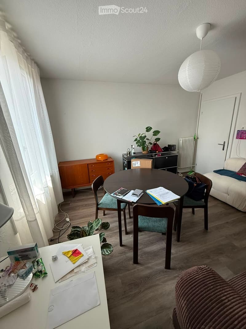 A modern, well-lit living space with a round wooden dining table, four chairs, a sofa, a wooden cabinet, a floor lamp, and a small desk with various items.