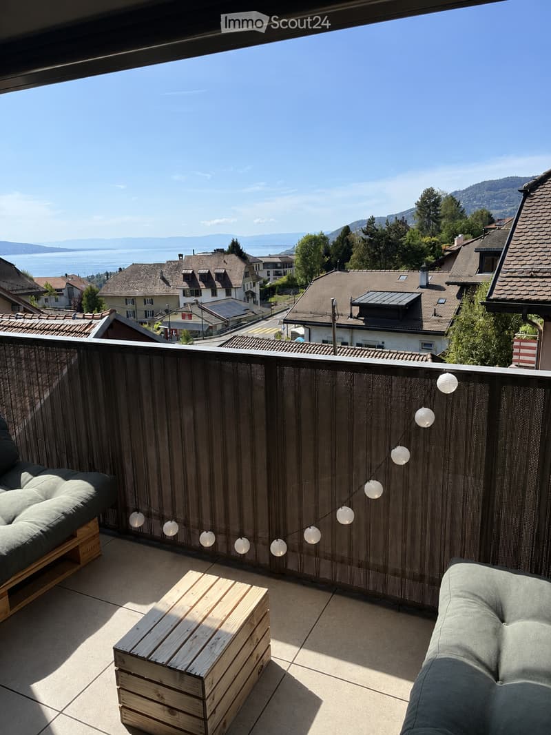 Balcony with a scenic view of a lake and mountains, featuring a wooden table and two cushioned seating areas.