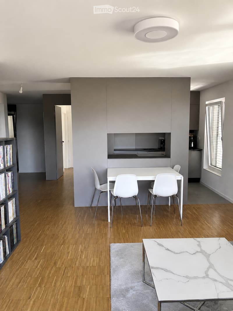 Modern, open-plan living and dining area with wooden flooring, white dining table with four chairs, a white coffee table with a marble top, a grey rug, and a bookshelf with books.