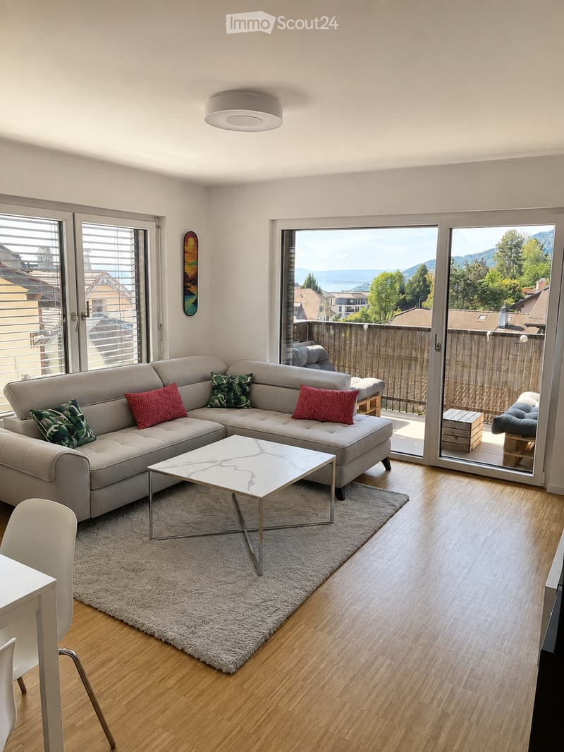 Modern living room with large windows offering a scenic view, featuring a grey sectional sofa, a white coffee table, a plush rug, and a dining area with a white table and chairs.