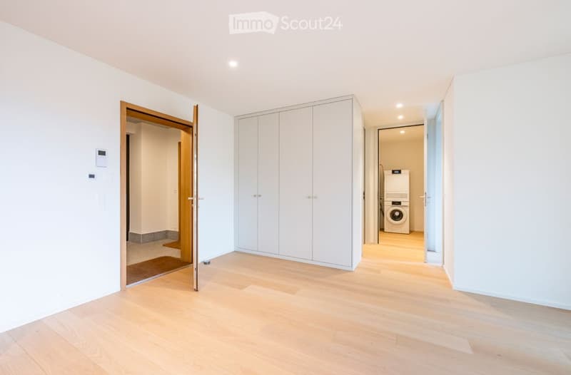 Empty room with light wooden flooring, white walls, built-in white closet, and an open doorway leading to a laundry area with a washing machine.