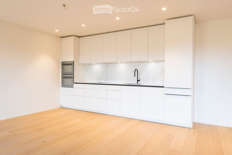 Modern, minimalist kitchen with white cabinets, a stainless steel oven, a sink with a modern faucet, and a refrigerator. The floor is light wooden, and the walls are white.