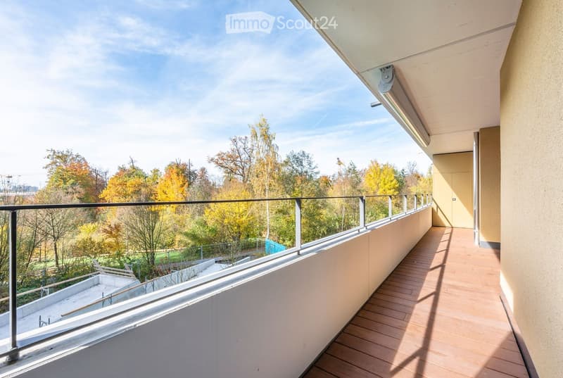 Balcony with wooden flooring, glass railing, and scenic view of autumn trees and a park area.