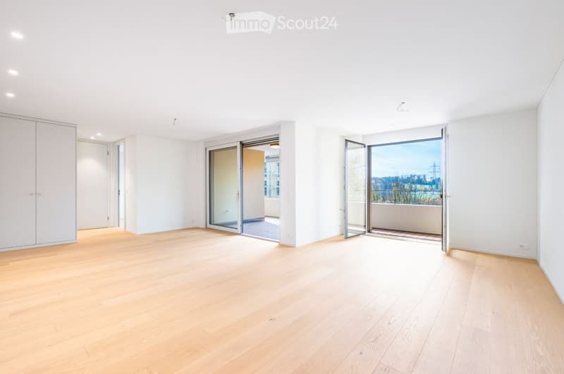 Bright, empty room with light wooden flooring, white walls, and large sliding glass doors leading to a balcony with a scenic view.