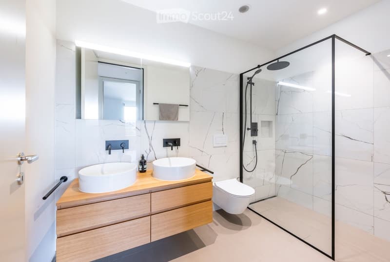 Modern bathroom with white marble tiles, double sink vanity, toilet, glass-enclosed shower, and a large mirror.