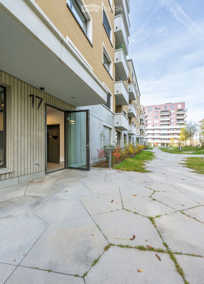 Modern residential building entrance with a glass door, concrete pathway, and small garden area with plants and flowers.