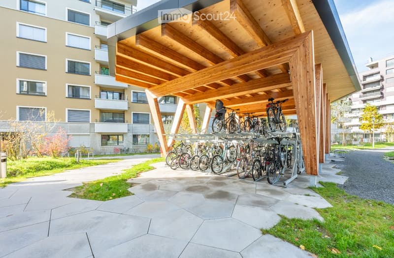 Outdoor bike parking area with wooden shelter, multiple bicycles, and modern building in the background.