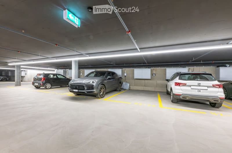 Underground parking area with several parked cars, marked parking spaces, and an emergency exit sign.
