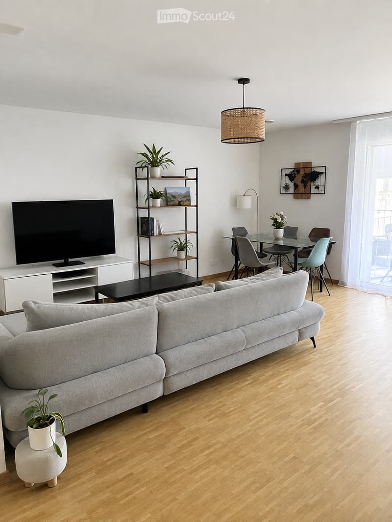 Modern living room with light wooden flooring, grey sectional sofa, coffee table, TV, shelving unit with plants and books, dining area with glass table and chairs, and a hanging woven pendant light.