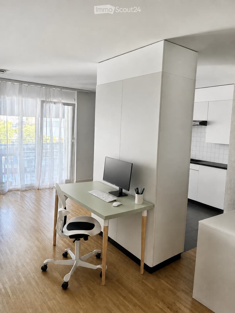 A modern, minimalist office space with a light green desk, ergonomic chair, computer monitor, keyboard, mouse, and a cup on the desk. The room features light wooden flooring and white cabinetry.