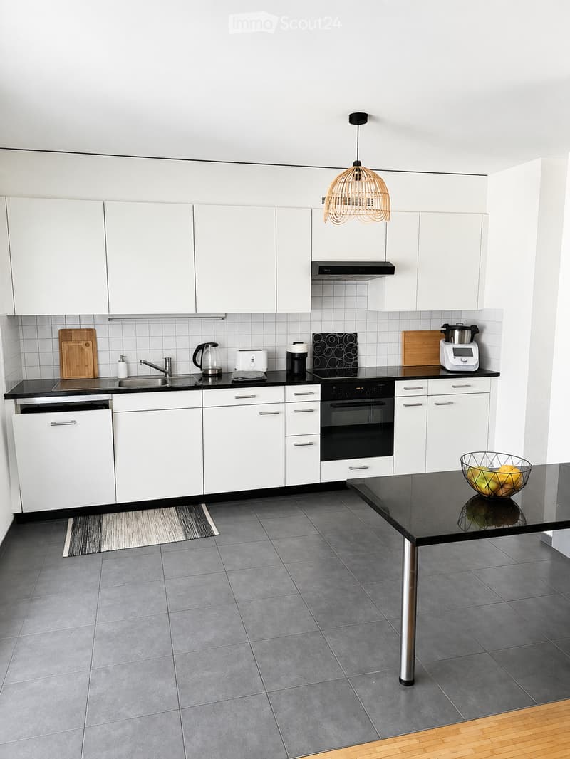 Modern kitchen with white cabinets, black countertops, stainless steel appliances, and a central island with a bowl of fruit.