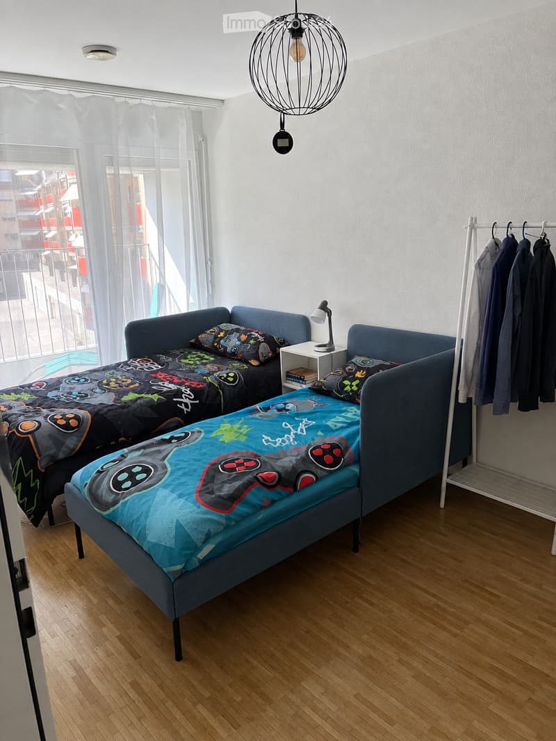 Cozy bedroom with two single beds, colorful gaming-themed bedding, a nightstand with a lamp, and a clothing hanger with jackets.