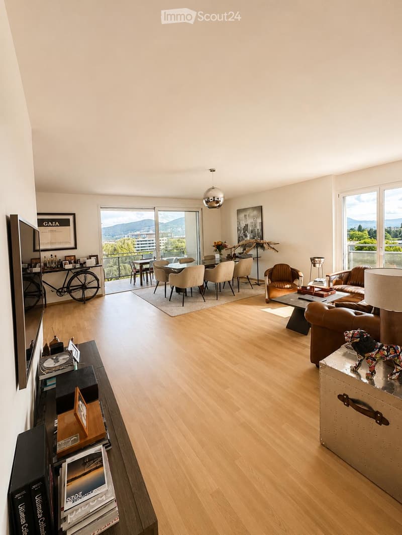 Modern living room with hardwood floors, large windows, and a balcony. Features a sofa, coffee table, dining area with chairs and table, and decorative items including a bicycle and artwork.