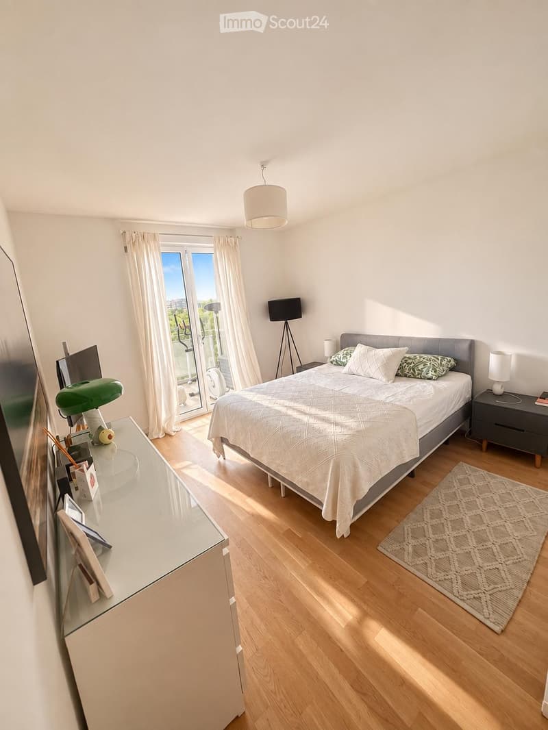 Bright bedroom with wooden flooring, large window with balcony access, bed with white bedding, nightstand, lamp, and a small table with a green lamp.