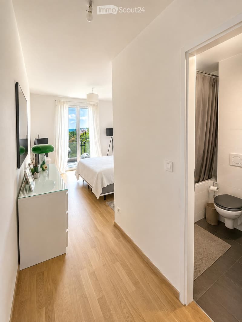 Bright bedroom with wooden flooring, large window with a view, bed with white bedding, nightstand with lamp, mirror, and a small table with decorative items. Adjacent bathroom with toilet and tiled floor.