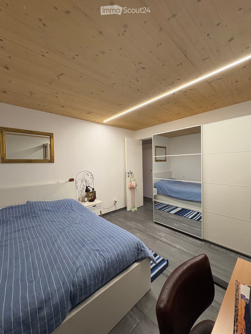 Cozy bedroom with a large bed, a chair, a small table, a mirror, a lamp, and a built-in wardrobe with a bunk bed inside.