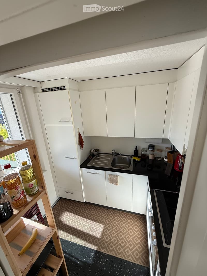 Modern kitchen with white cabinets, black countertops, and a stainless steel sink. Visible items include a coffee maker, bottles, and a small shelf with food items.