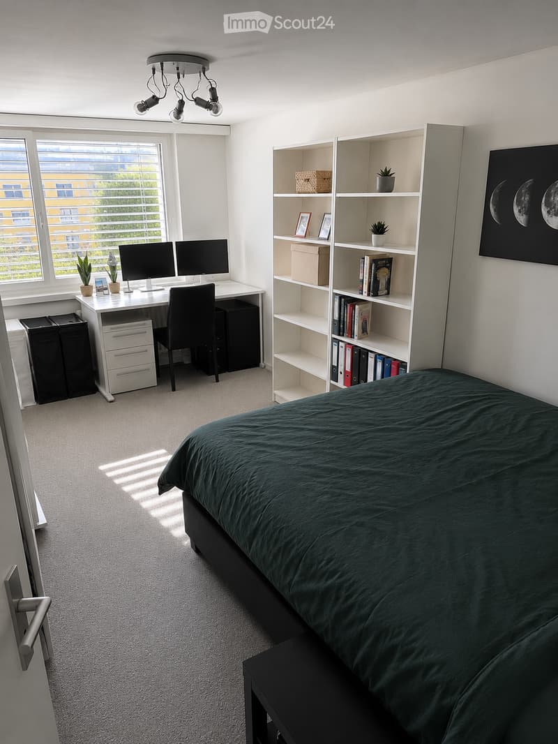 Modern bedroom with a large bed, desk with two monitors, bookshelf, and potted plants. The room features white walls, carpeted flooring, and large windows with blinds.