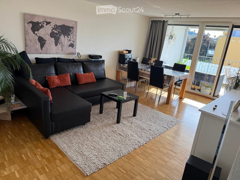 Modern living room with a black sectional sofa, coffee table, dining area, and large windows leading to a balcony.