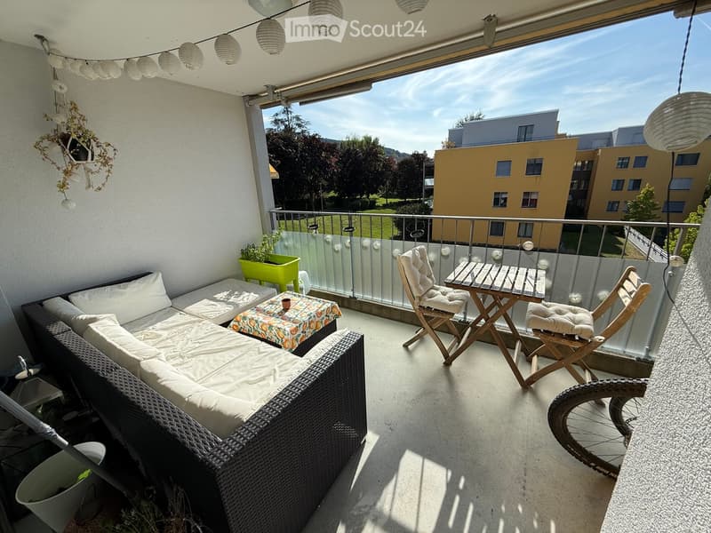 Balcony with a white sofa, small table, two wooden chairs, and a bicycle wheel. The balcony has a glass railing and a view of a park and buildings.