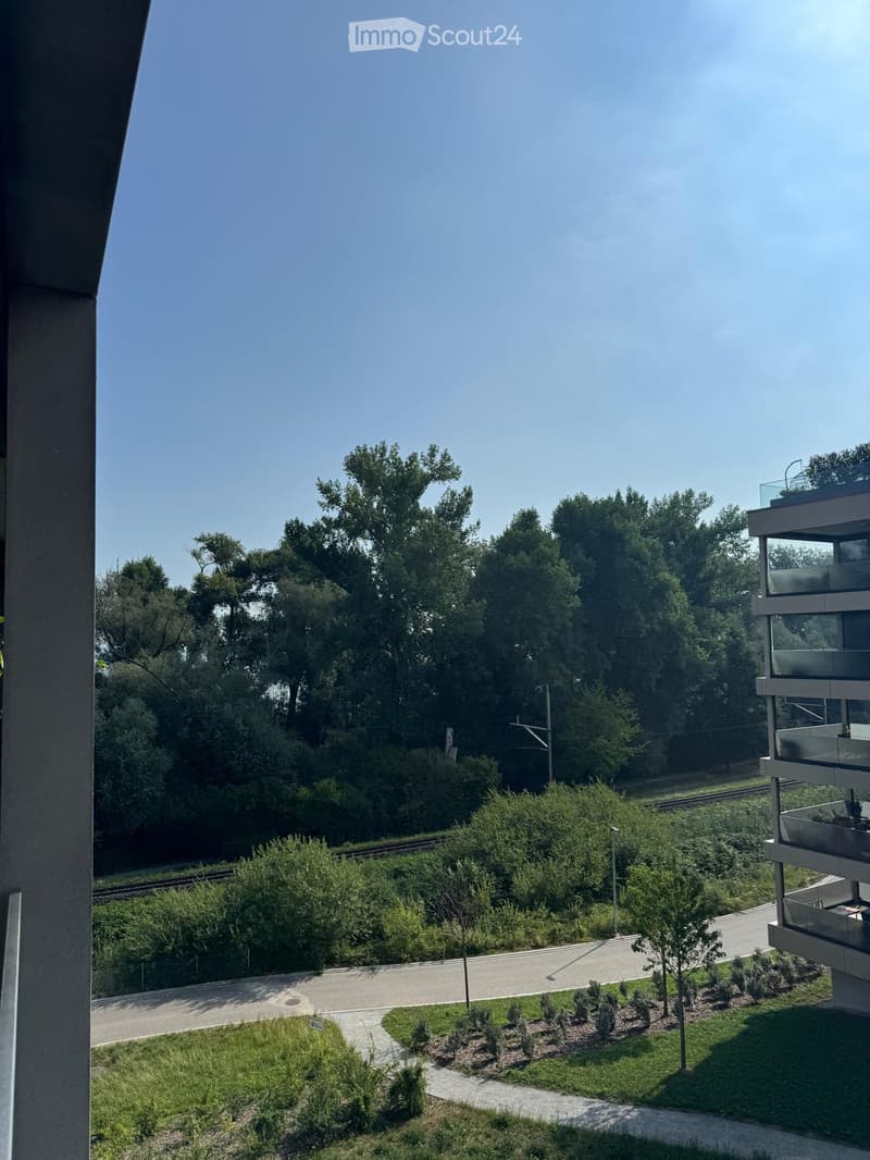 Balcony view overlooking a landscaped area with trees, a pathway, and a modern building with glass railings.