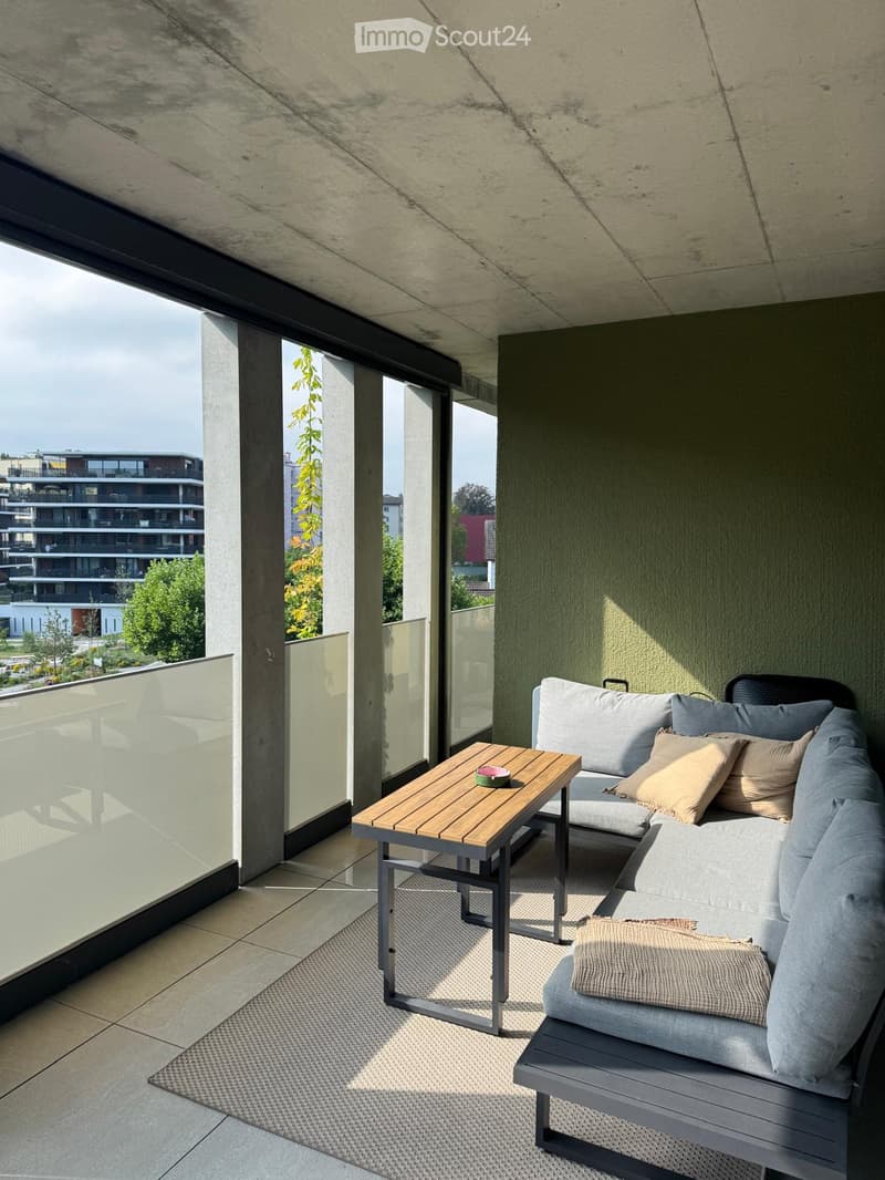 Modern balcony with a grey sofa, wooden table, and decorative pillows, featuring a view of the city and greenery.