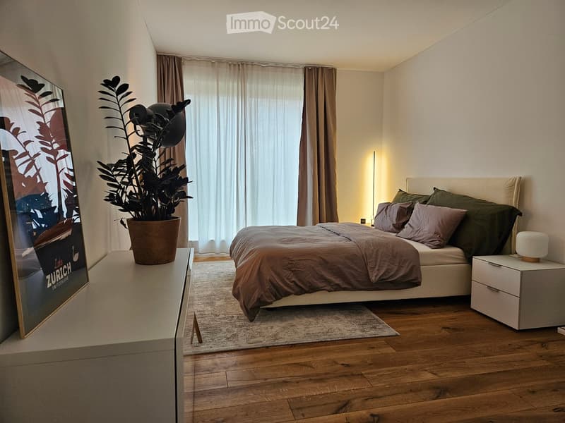 Modern bedroom with wooden flooring, a large bed with pillows and a duvet, a nightstand with a lamp, a plant on a side table, and a large picture frame on the wall.