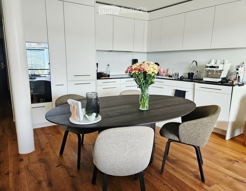Modern kitchen with white cabinets, a round wooden dining table, four chairs, a vase with flowers, and a coffee machine on the counter.