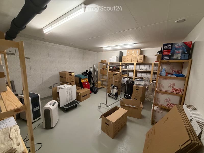 Empty storage room with concrete walls, fluorescent lighting, and various boxes and storage items scattered throughout.
