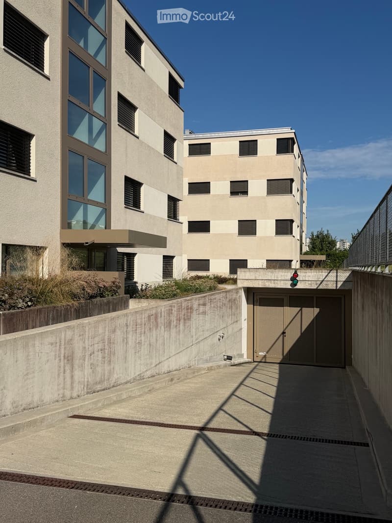 Modern multi-story building with large windows and a raised ground floor entrance, featuring a concrete ramp and garage access.