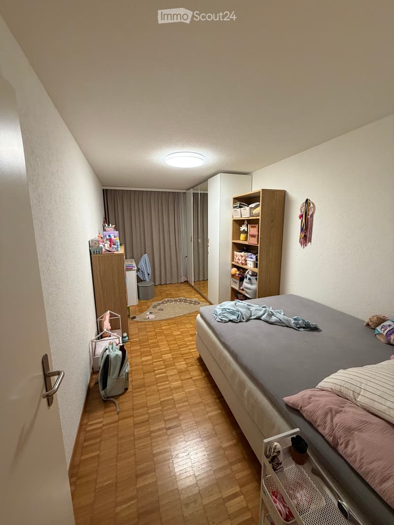 A small, well-lit bedroom with a single bed, a backpack, a shelf, a mirror, and a curtain. The room features wooden flooring and a closet with a mirror.