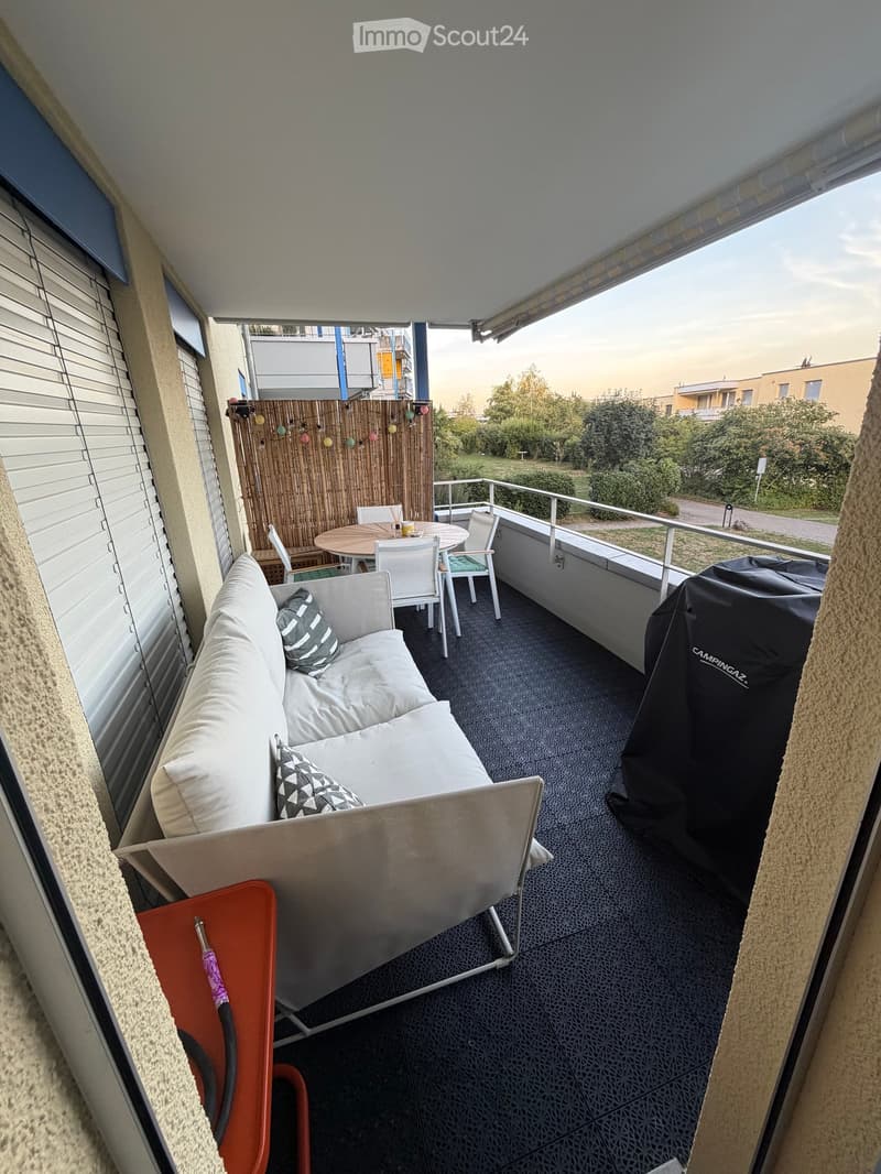 Balcony with a sofa, table, chairs, and an umbrella, featuring a view of greenery and a covered outdoor area.