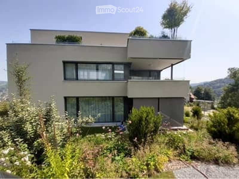 Modern two-story house with flat roof, large windows, and balcony on the upper floor. The house is surrounded by a well-maintained garden with various plants and shrubs.