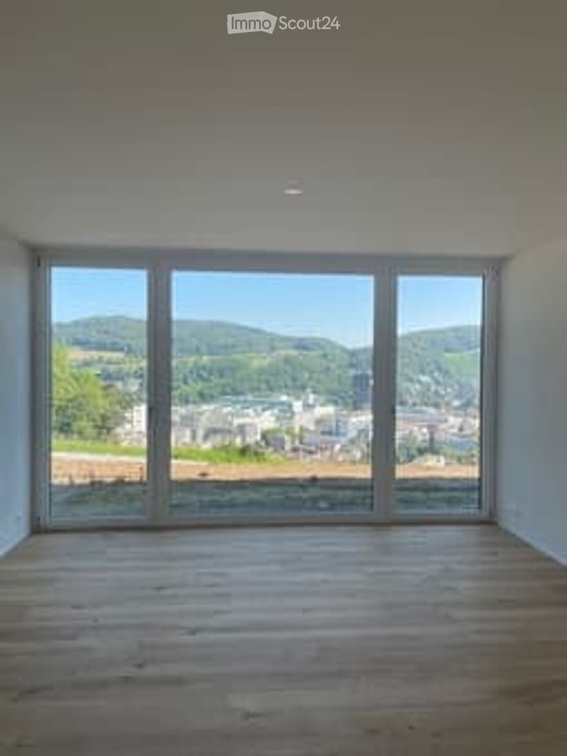 Completely empty room with large windows offering a scenic view of hills and a town. Wooden floor, white walls, and no furniture.