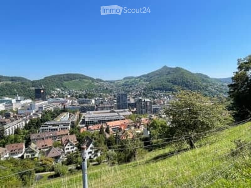 Panoramic view of a cityscape with buildings, hills, and green fields under a clear blue sky.