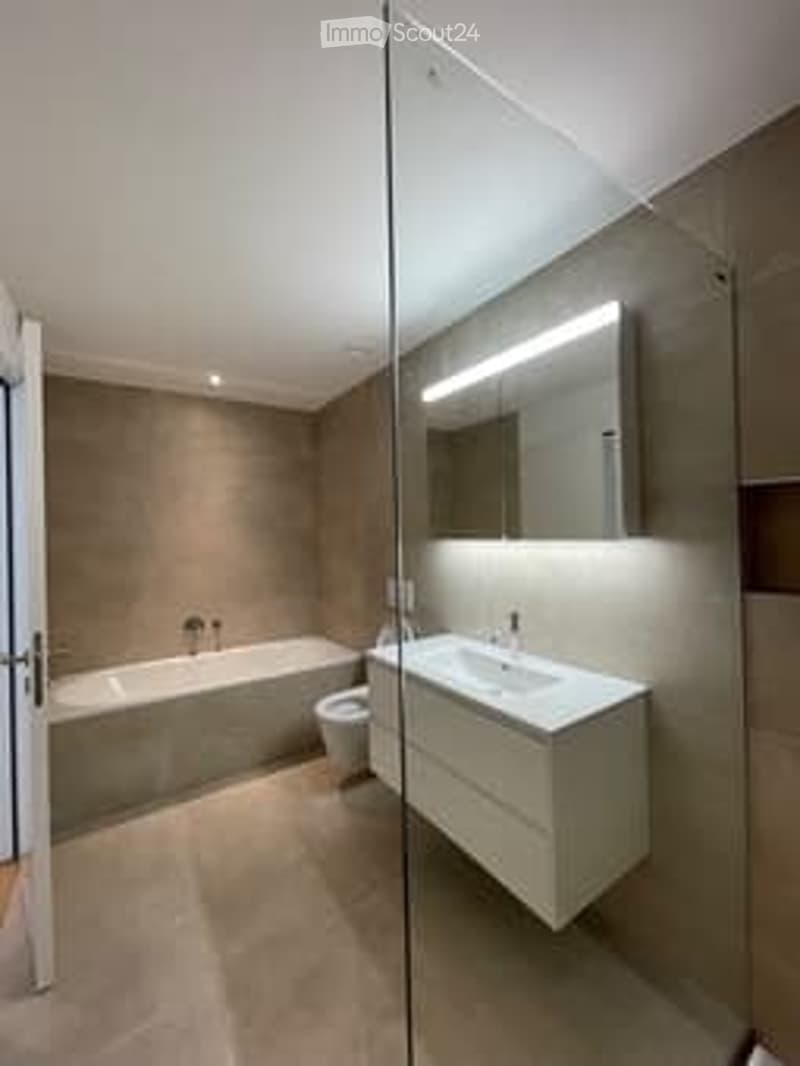 Modern bathroom with a large bathtub, a sleek sink, and a mirrored vanity. The space features warm beige tones and a glass partition.