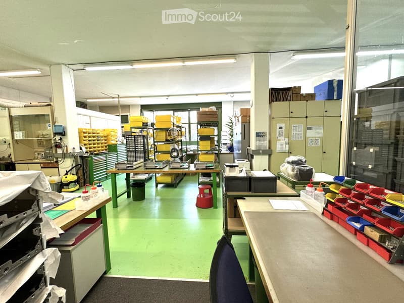 A workshop with green floor, multiple tables, shelves, and storage boxes, a red stool, and a blue chair.