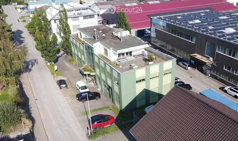 Aerial view of a building with multiple floors, green roof, and parking area with several cars parked.