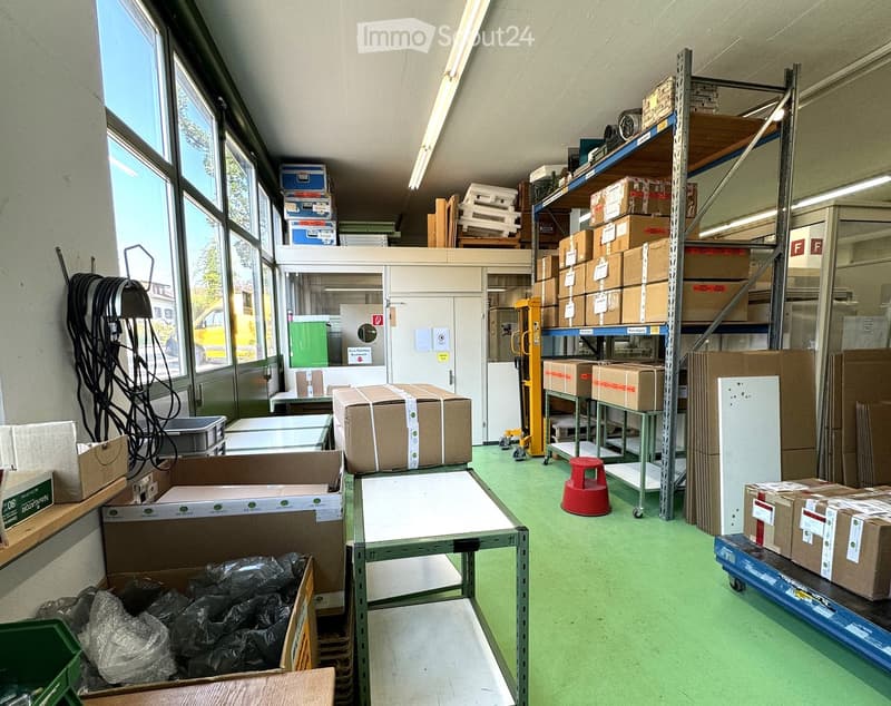 Warehouse with large windows, green floor, shelves with boxes, trolleys, and storage boxes